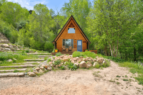 A cozy wooden cabin surrounded by greenery and rocks, with a clear blue sky in the background.